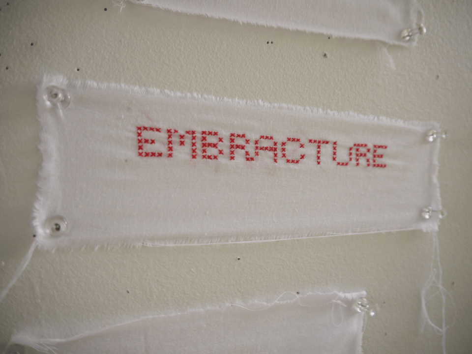 the capitalized word "embracture" cross stitched in red onto a strip of light white cloth