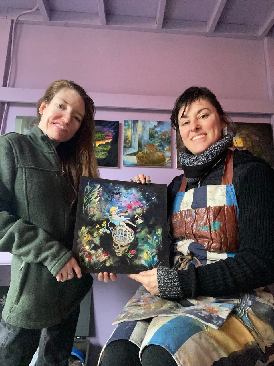 Two women standing together, one on the left with reddish-brown hair wearing a dark green jacket, author Taylor Yount, and a woman on the right with dark brown hair wearing a multicolored apron and a black sweater, illustrator Veta Bakhtina, are both holding one of the illustrations from the book, My Sutured Mind, featured from the poem “Perceptual Limbo.”