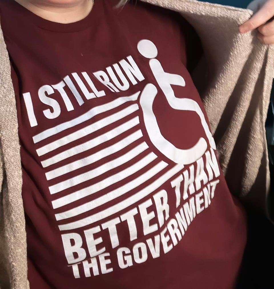 Yaakov from the neck down, wearing a beige cardigan over a maroon tee that has the wheelchair symbol for disability and reads "I still run better than the government"