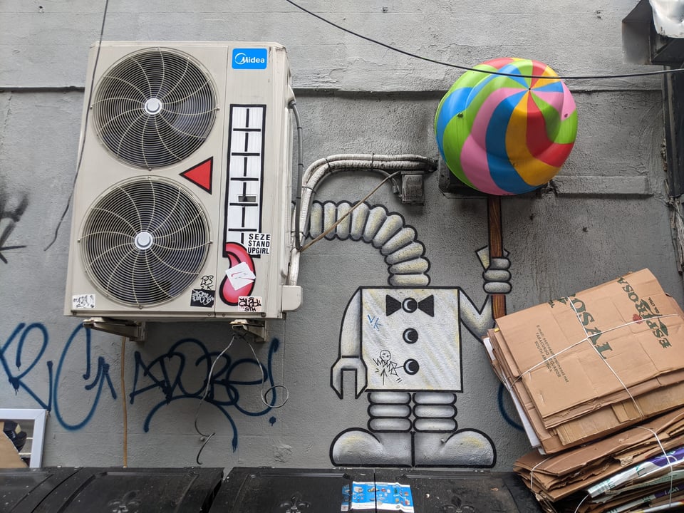 An air conditioner painted to look like a robot with a tiny bow tie, holding a swirly colorful popsickle.