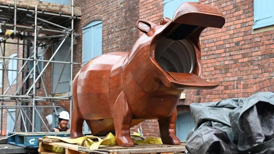 A chonky wooden hippo standing on pallet. Its mouth is wide open, revealing there is a large hole. The belly touches the floor, suggesting the slide emerges from its bottom. It is glossy, and beautiful and a workman in a hardhat is standing near it and grinning.