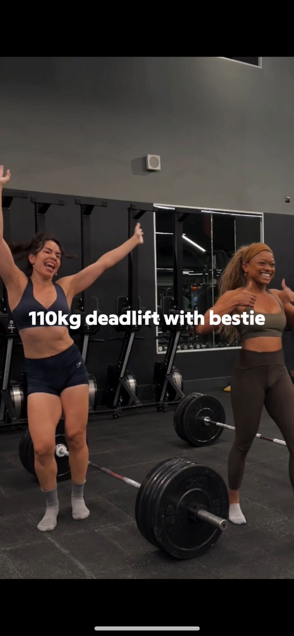 In a gym, two women celebrate after deadlifting. Jacinda, on the left, has her arms raised in excitement. I stand on the right, smiling and giving a thumbs-up. Two loaded barbells lie on the floor in front of you both. The text “110kg deadlift with bestie” appears across the center of the image.