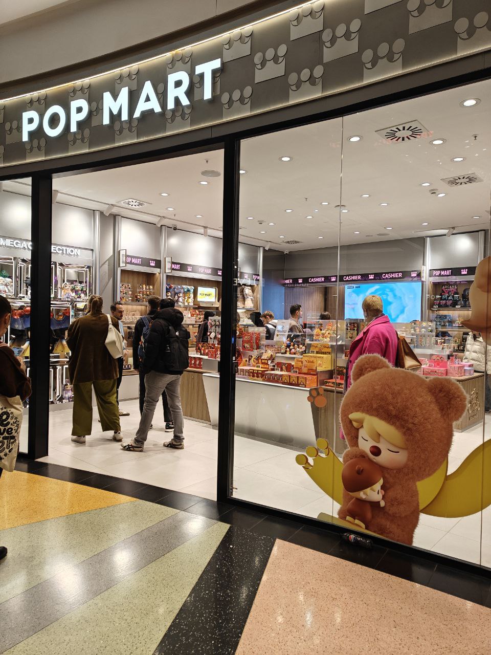 A photo of popmart, in the Alexa center mall. It's brightly lit with overhead flourescent lights and a small cluster of people just inside the door. On the glass of the window is a large vinyl print of one of the blindbox characters