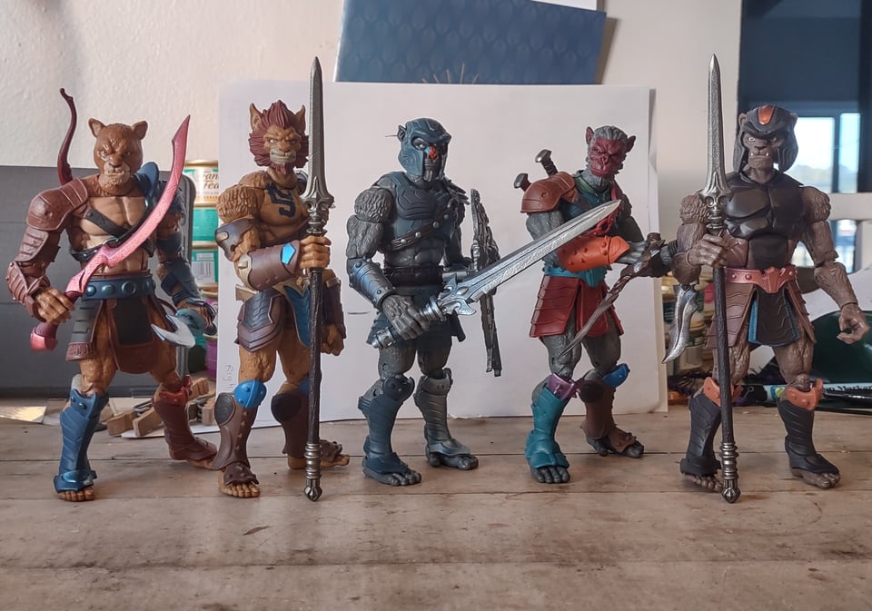 Five action figures of animal-men in fantasy armor stand on a wooden tabletop. They hold spears, swords, and daggers in their hands.