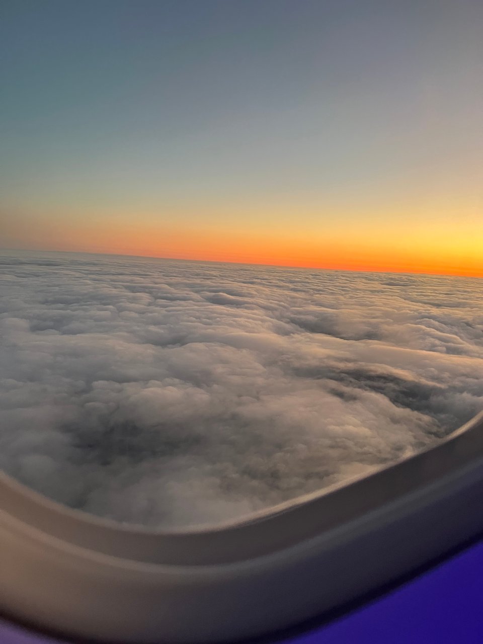 Looking out of a plane window at grey clouds and a bright orange sunset on the horizon.