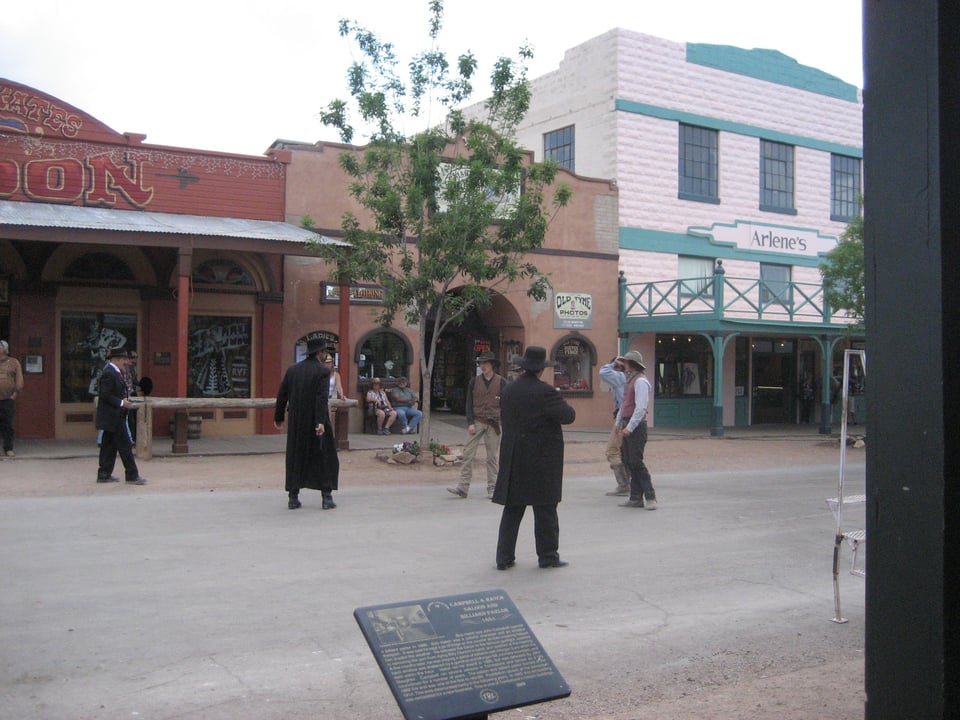 A group of cowboys in a standoff in the street...there are three buildings that face onto a classic boardwalk, one red, one light brown, and one blue and white.