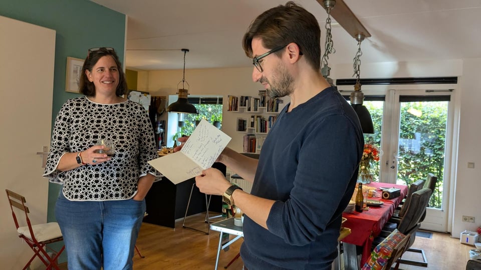 A man reads a birthday card he is holding while a woman holding a glass looks on smiling.