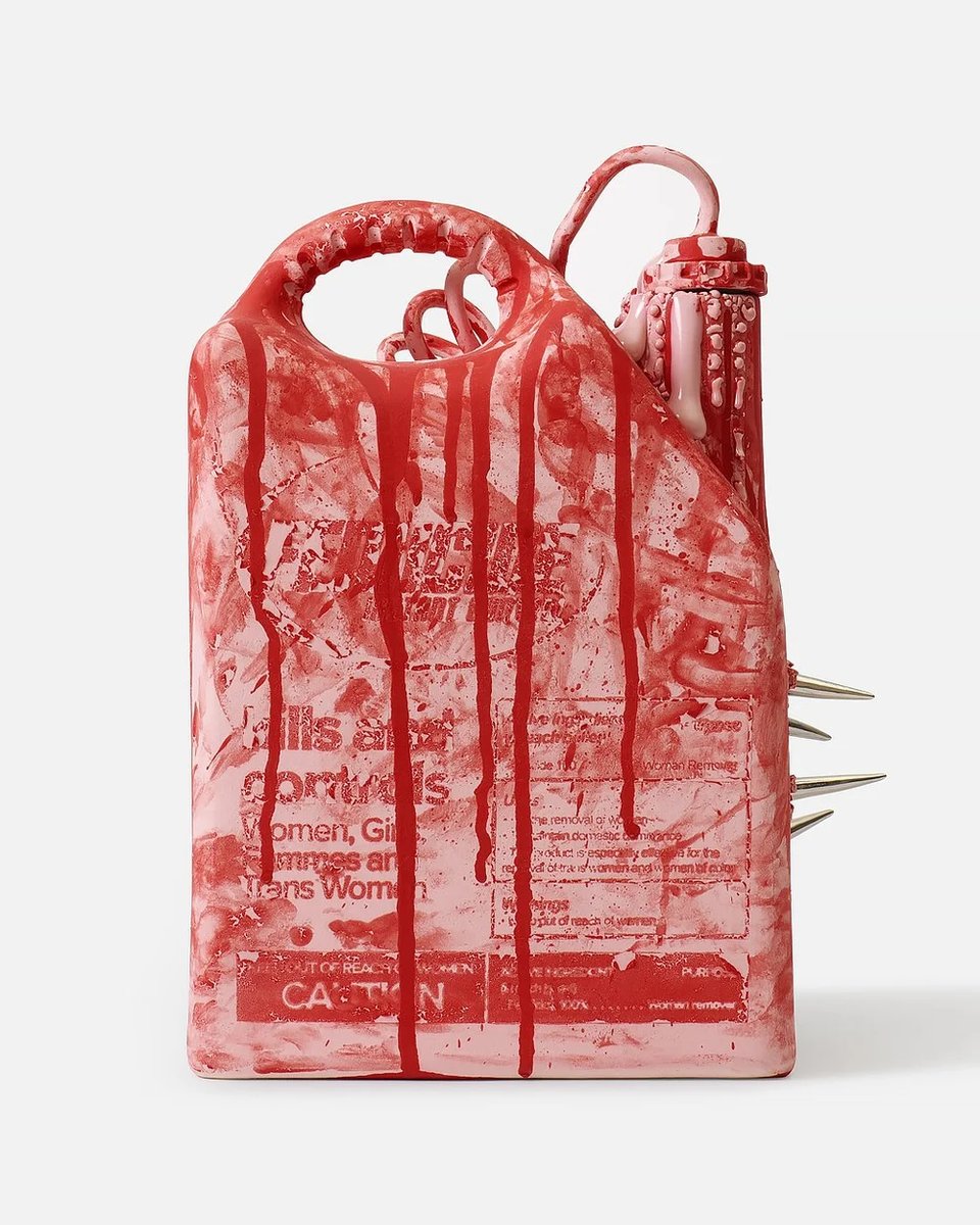 A sculptural ceramic artwork resembling a plastic jug or chemical container, glazed in mottled pink and blood-red with drips running down its face, bearing pesticide-style warning text reading "kills and controls Women, Girls, Femmes and Trans Women" and "CAUTION," with a looped handle at top, a capped spout on the upper right, and three metal spikes protruding from its right side, photographed against a plain white background.