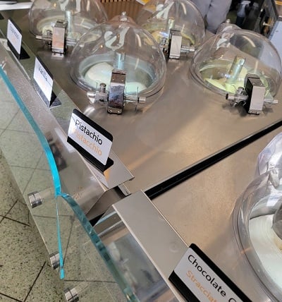 Gelato machines embedded in a stainless steel counter. There are signs indicating the flavors including pistacchio and stracciatella