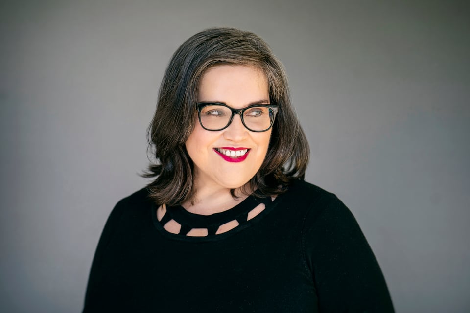 A photo of me, a woman in a black sweater, looking off to the side, wearing red lipstick and glasses with dark frames, smiling