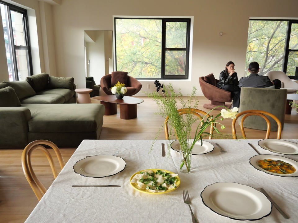 [Delicious looking plates of food sit beside place settings before a communal lunch. In the background, two people sit in comfy chairs before big windows overlooking the street.]