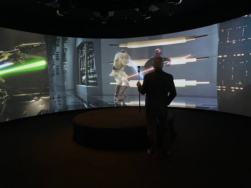 A man stands in front of a screen showing three different lightsaber fights