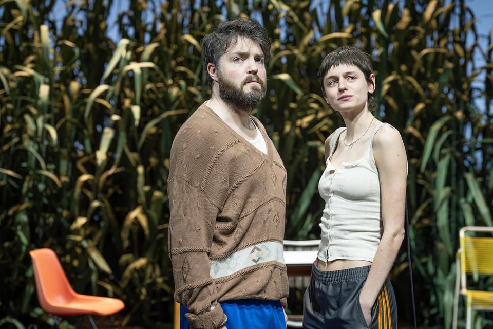 Tom Burke and Emma Corrin in Thomas Ostermeier and Duncan Macmillan's The Seagull. Burke has black hair and a big black beard, is wearing a loose brown patterned jersey and blue gym shorts; Corrin has short black hair, is wearing a beige buttoned tank top and black and yellow-striped sweatpants. They are standing in front of a cornfield and some plastic chairs.