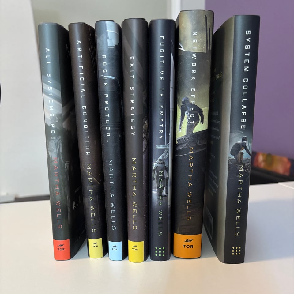 All the hardcover copies of The Murderbot Diaries by Martha Wells, spines out. Two of the books have different spine designs and one of the books is obviously taller.