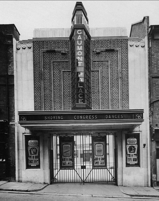 A brick and stone entrance arch with closed wrought iron gates. The brickwork has neat fins in it, and the stone has stylised waves at the top. A vertical fin rises from the projecting canopy to the roofline, and is a glass and metal illuminated sign for the "Gaumont Palace".