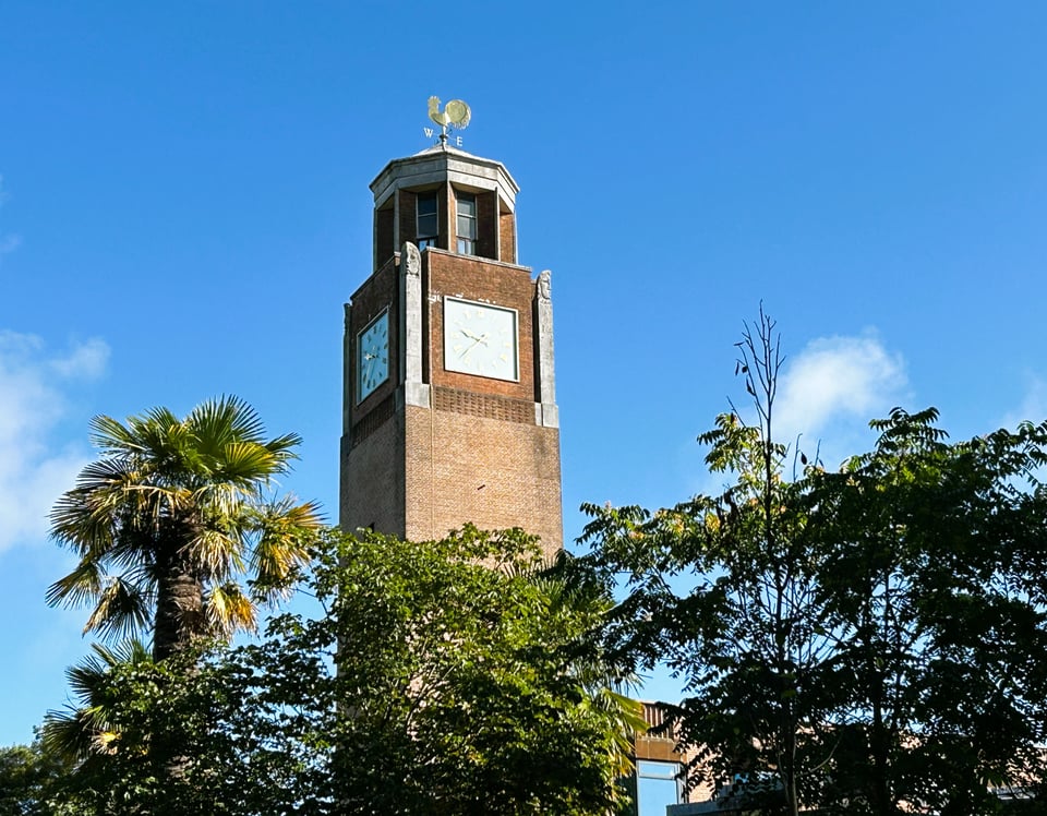 A plain square brick tower rises from a mass of trees including palm trees. At the top, two of the four square clock faces are visible, and the brickwork steps in to make space for four slender obeslisks that have heads carved on the tops of them. Not at all creepy. Above a parapet, an octagonal turret made mostly of windows rises to a stone roof with a golden weathervane on it. The wind sits in the west.