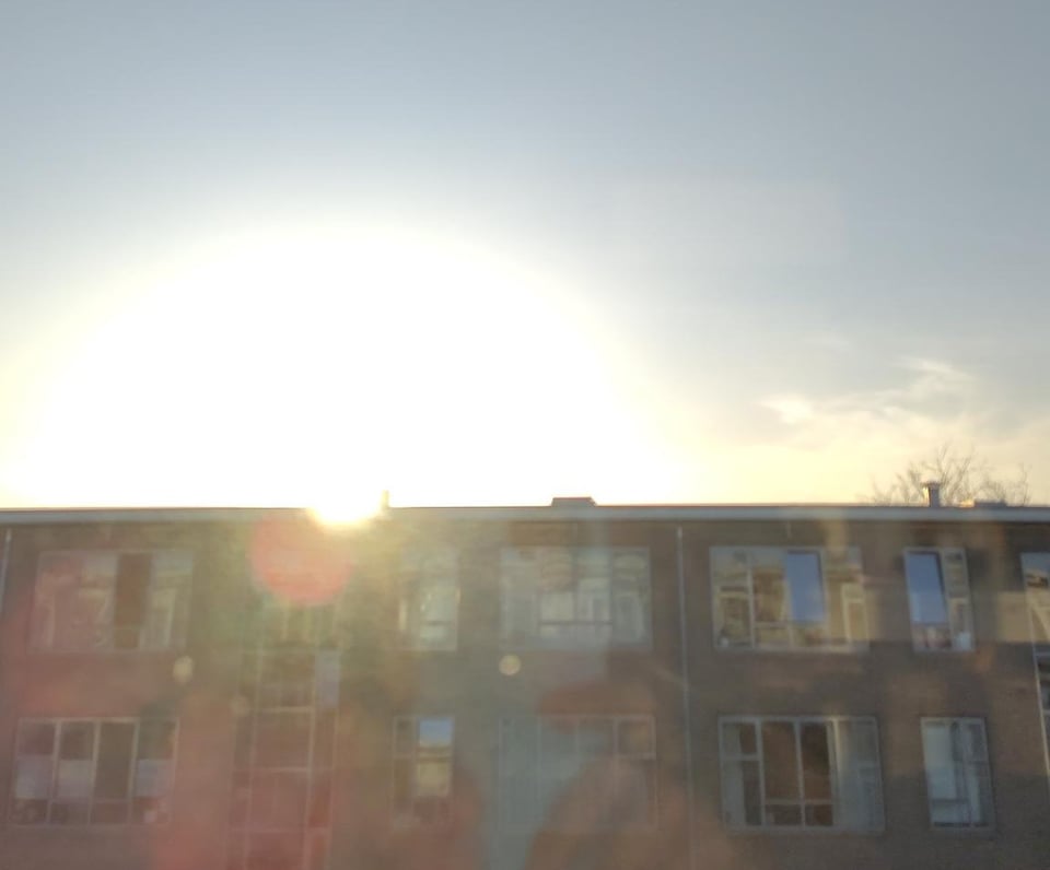 Image description: a phone picture of the sun over a row of buildings. the brightness has washed out the area around the sun, which sits in an almost cloudless blue sky. the reflections of my hands holding a blue phone can be faintly seen. end ID.