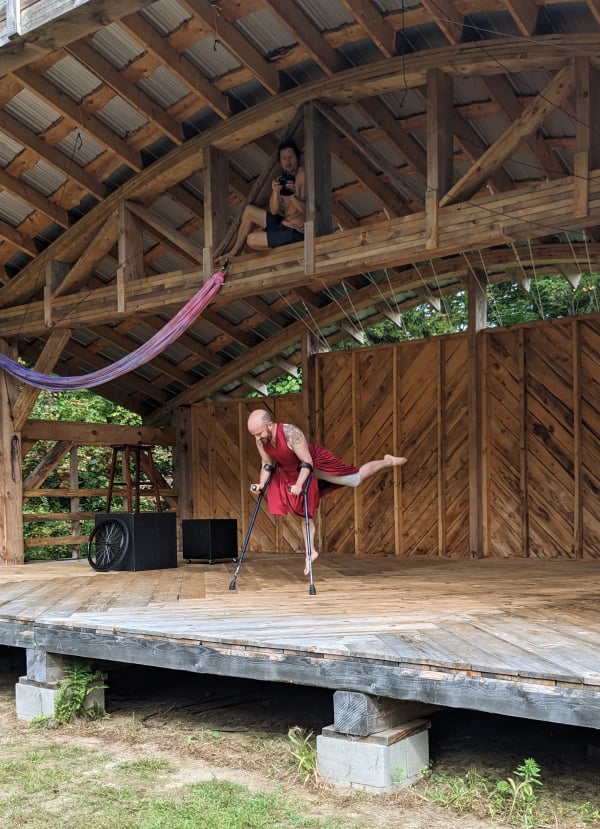 I am mid-leap on my crutches on a natural wood stage, with herringbone floor and back walls, a curved ceiling, and heavy cross beams. Videographer Andreas is perched on one beam directly above me, filming down. I'm in my red wrap dress, which floats in the air along with one leg kicked up back. The set pieces are assembled behind me.