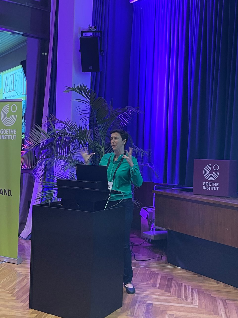 A woman stands at a podium gesturing with her hands. The Goethe Institute logo sits on the stage behind her.
