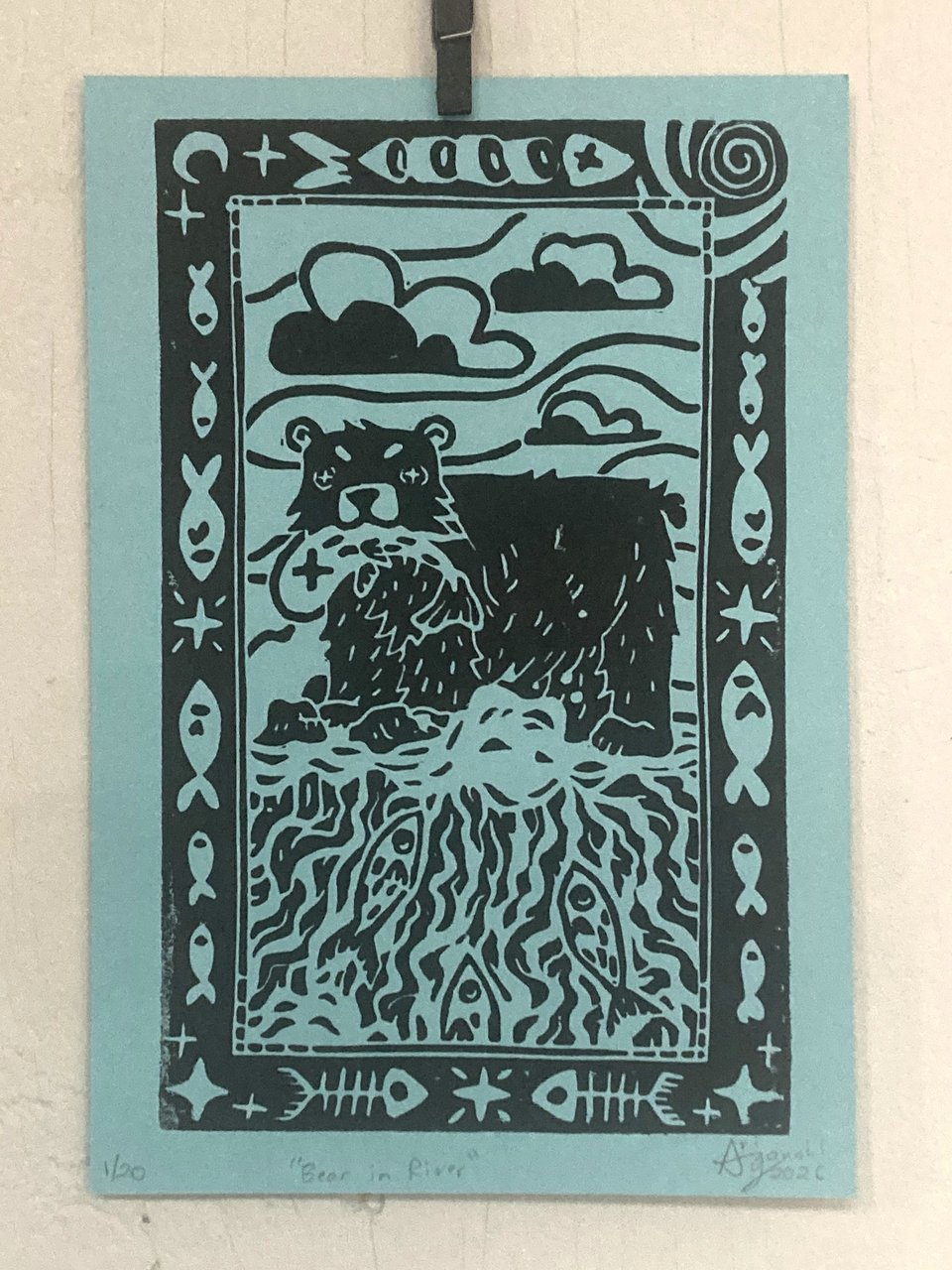 Relief printing with black ink on blue paper of a bear standing at the top of a waterfall with a fish in its mouth