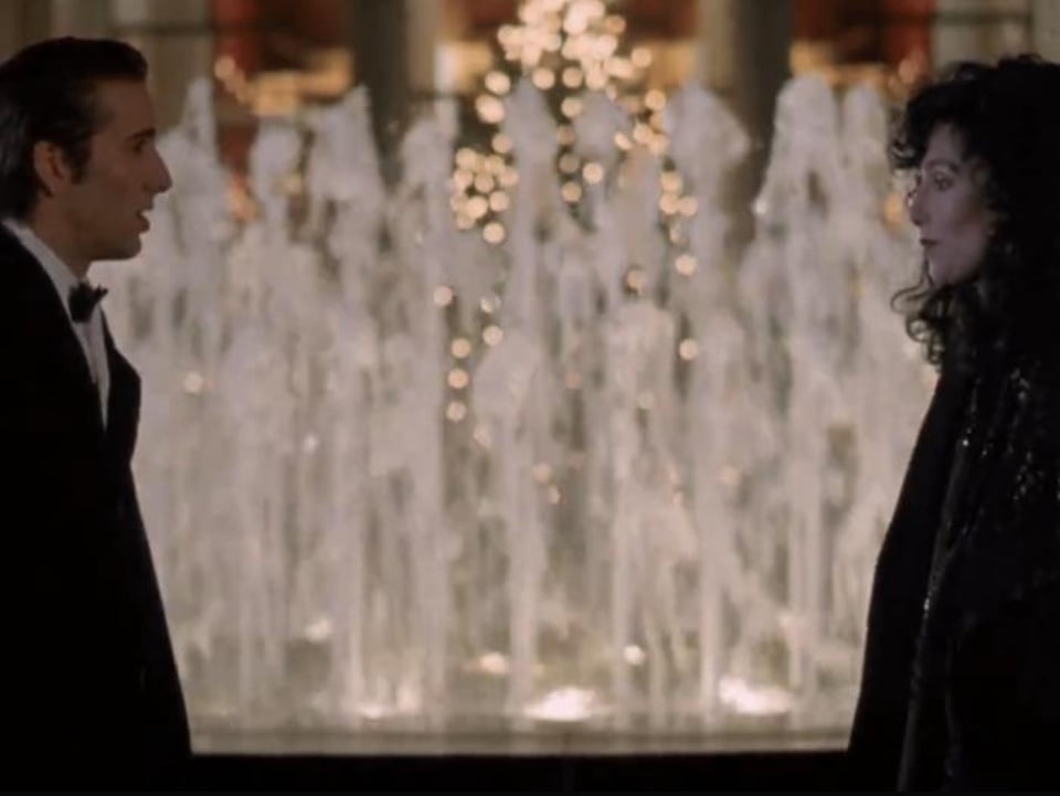 Nicolas Cage and Cher as Ronny and Loretta in Moonstruck. Both are wearing formal attire and stand facing each other in front of the Lincoln Center fountain with the Metropolitan Opera House in the background.