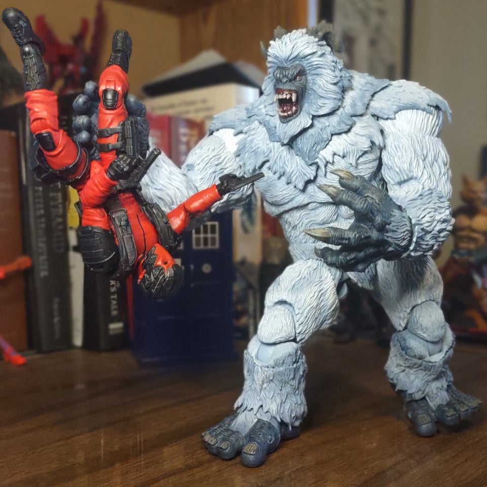 An huge action figure of a white-furred Yeti stands on a wooden desk. His mouth is open in a snarl showing lots of teeth, and he has curling horns and huge claws. Dangling upside down from one of his hands is a vaguely militaristic figure of a man in a red and black uniform, half the Yeti's size in every direction. The red soldier has two pistols out, but they're probably not going to do him any good.