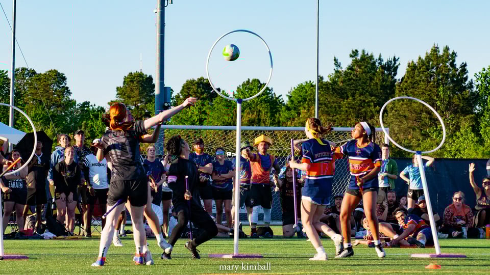 Creighton chaser Mimi Baldwin with the championship winning goal during the D-I finals against Virginia at USQ Cup 2025. The ball is framed in the center hoop and there's a man wearing a cheese hat in the background looking amazed.