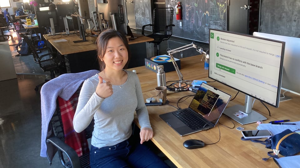 Katherine sitting at her desk giving a thumbs up while looking at the camera with her monitor showing a merge PR screen on the desk next to her.
