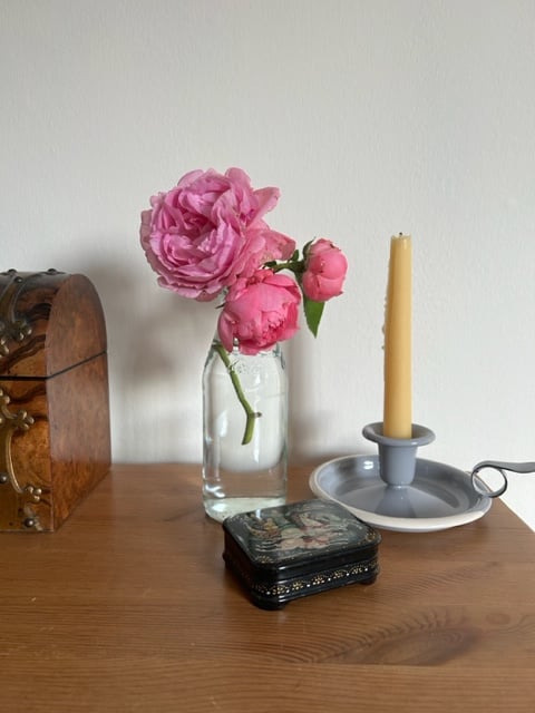 A small cluster of three pink roses in a glass bottle, next to a tall beeswax candle and a small wooden box