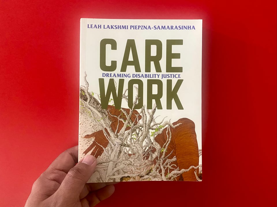 A hand holding a copy of the 2018 book Care Work Dreaming Disability Justice by Leah Lakshmi Piepzna-Samarasinha against a red tabletop background.