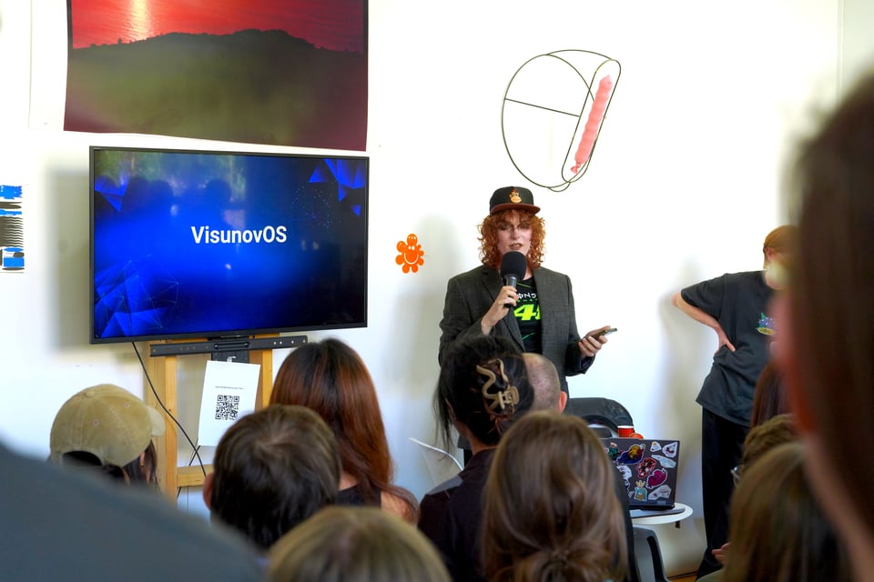 A presenter - in classic "E3 keynote" outfit - gestures in front of an audience. The screen next to her says "VisunovOS"