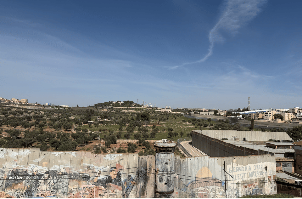 A photograph taken from a high point, showing, in the foreground, the same concrete separation barrier with graffiti that is shown in the previous photo. On the other side of the wall are green areas and olive trees. In the distance, what looks like small towns consisting of clusters of buildings