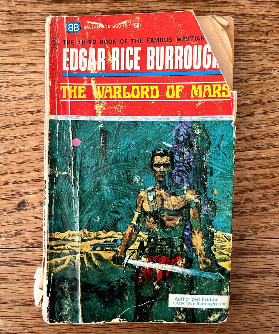 A battered paperback copy of The Warlord of Mars, by Edgar Rice Burroughs. The cover illustration depicts a shirtless man with a sword standing against an otherworldly background.
