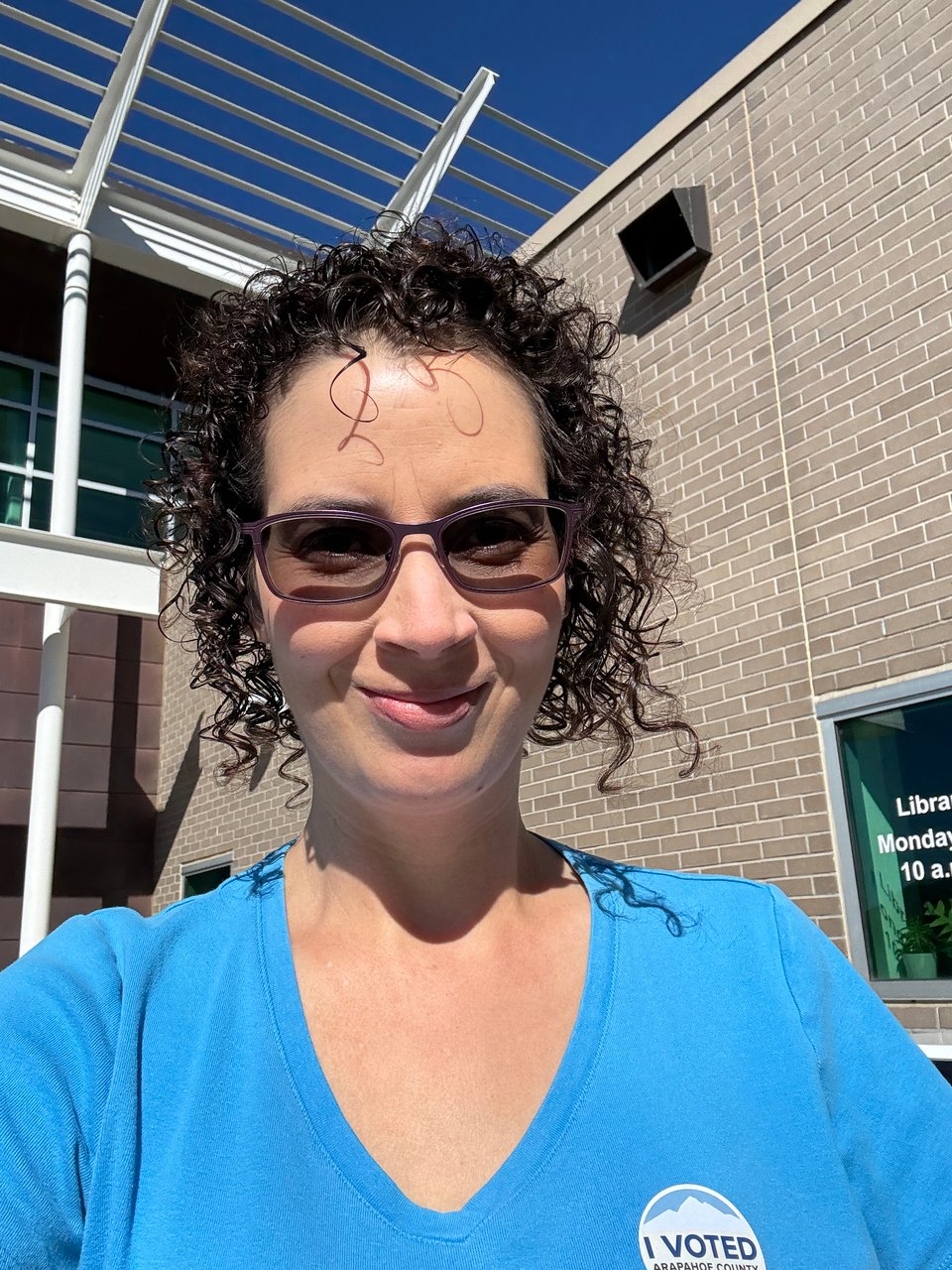 Amanda Helms in a blue shirt, outside a branch of Aurora Public Library, wearing a blue shirt and sporting her snazzy I Voted sticker