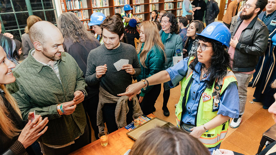 A woman in a blue hardhat and high-visibility vest pointing at party goers having a good time playing a game in a library.