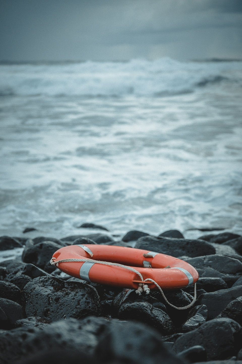 Photo of a bright orange life ring on a rock jetty in front of a roiling ocean that leads out to a grey, cloudy, sky.