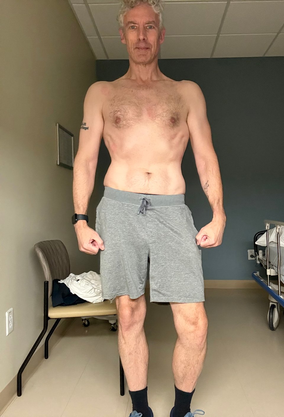 full-body photo of shirtless mid-50s white man in a pair of gray sweat-shorts and black socks. He's in a medical exam room, and his body looks decent for his age.