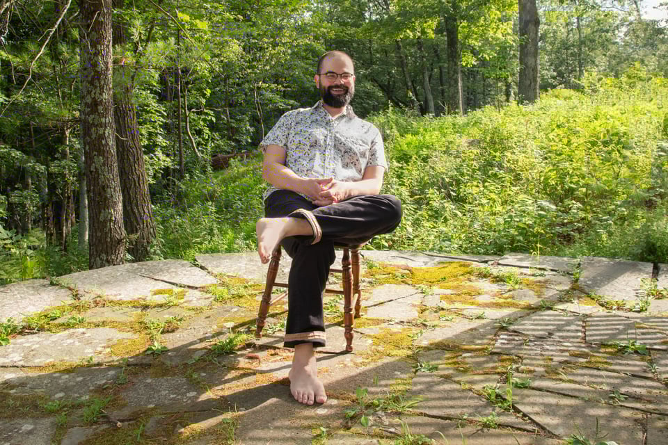 Gabriel sits reclined in a wooden chair on mossy flagstones surrounded by bright green foliage and sunlight. They are wearing black pants and a white short-sleeve button-down shirt covered in images of cats.