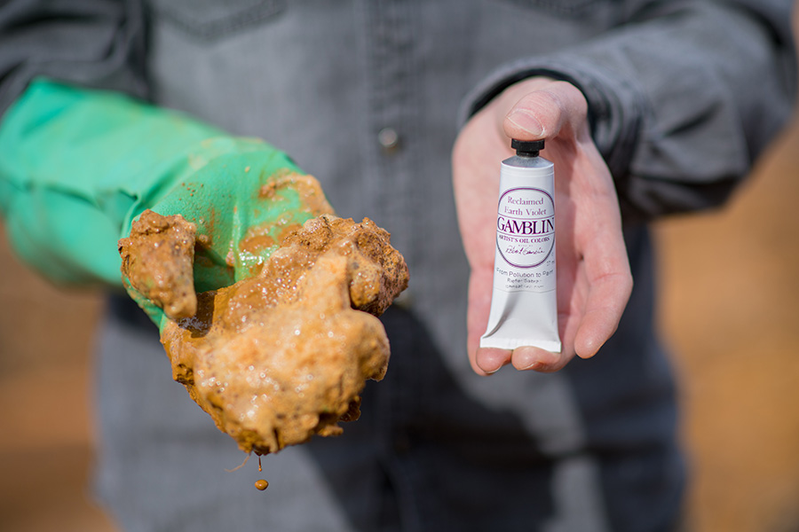 The photo of a man standing facing us with his hands stretched out towards us. His seft hand is covered in the thick plastic glove, and the fingers and palm are covered in a orange sludge. His left hand is bare and is holding a tube of paint, which is called "reclaimed earth violet"
