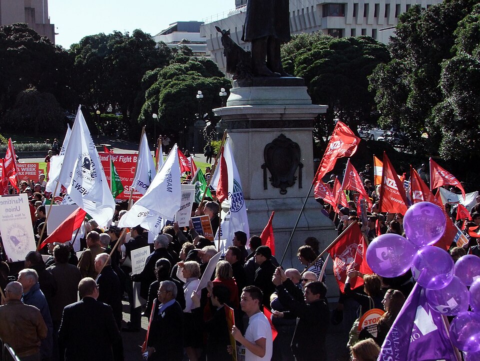 On the lawn of Aotearoa/New Zealand parliament grounds, unionists gather around the Seddon Statue. Flags include multiple NZEI flags, Maritime Union flags, and a Nurses Organisation Flag with matching purple balloons in the foreground. In the background, flags and banners of the now-defunct Service and Food Workers union are visible, along with a Green Party flag.
