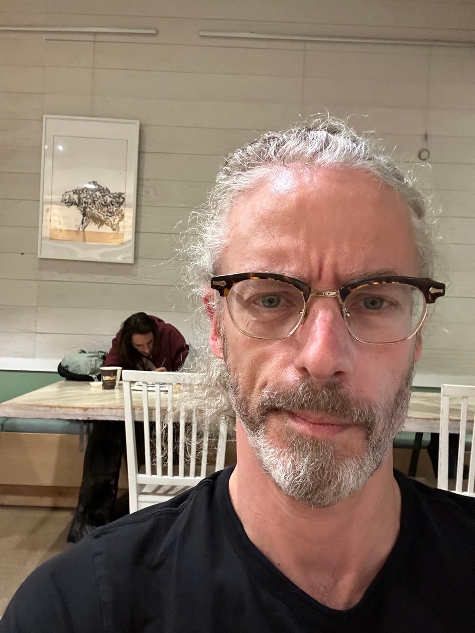 selfie of white man in black T-shirt & glasses in a coffee shop, with another man in the background, hunched over a table and painting a watercolor