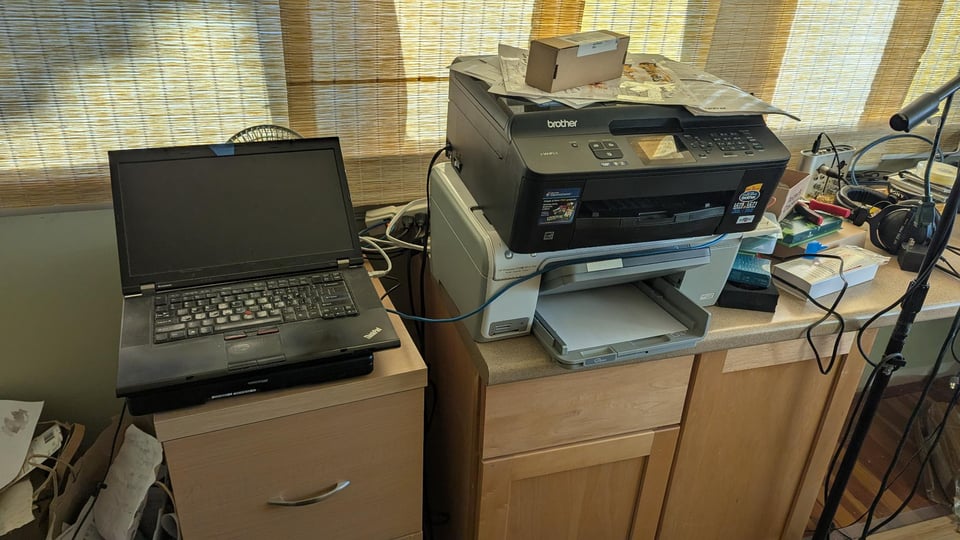 An ancient looking ThinkPad sits atop a filing cabinet, next to what appears to be two printers stacked one on top of the other.  A mess of half-completed electronics projects is just visible on a bench to the right of the printers.