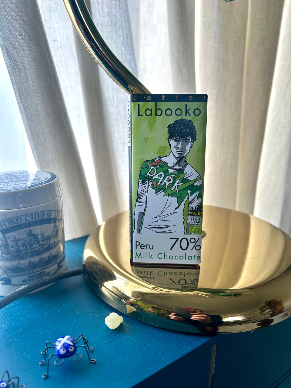 A bar of Zotter chocolate is propped against a brass reading lamp with cream curtains behind. There's a glass bug with a saltire flag on it and a bead of green frankincense. Image by Rowan Ambrose