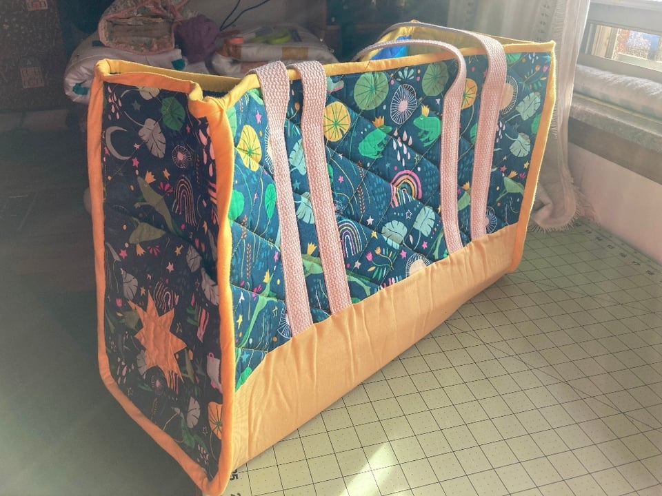 A quilted tote bag rests on a desk by a bright window. The body of the bag is split into two fabrics: one is a yellow fabric forming the bottom portion and the binding of the edges; the other is a green fabric with patterns of frog princes, lily pads, rainbows, rain drops, and rain boots. There are two sets of cotton webbing straps attached to the bag, and the side pocket shows a pieced together and quilted star block.
