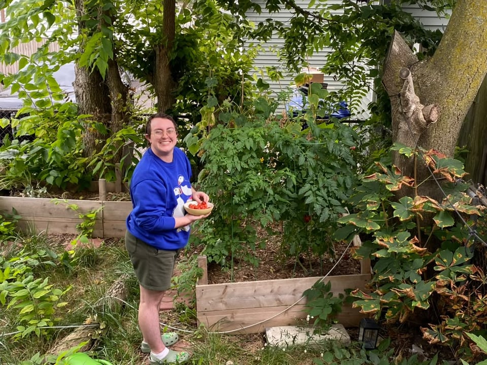 Emory smiling next to the raised garden beds where his tomatoes are growing. He is holding a bowl of harvested tomatoes.