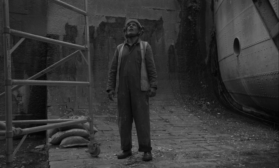 a man wearing a hi vis and overalls spatted with paint stands in the centre of frame looking up. To the left of frame is some scaffolding and on the right the edge of a ladder appears. The image is in black and white.