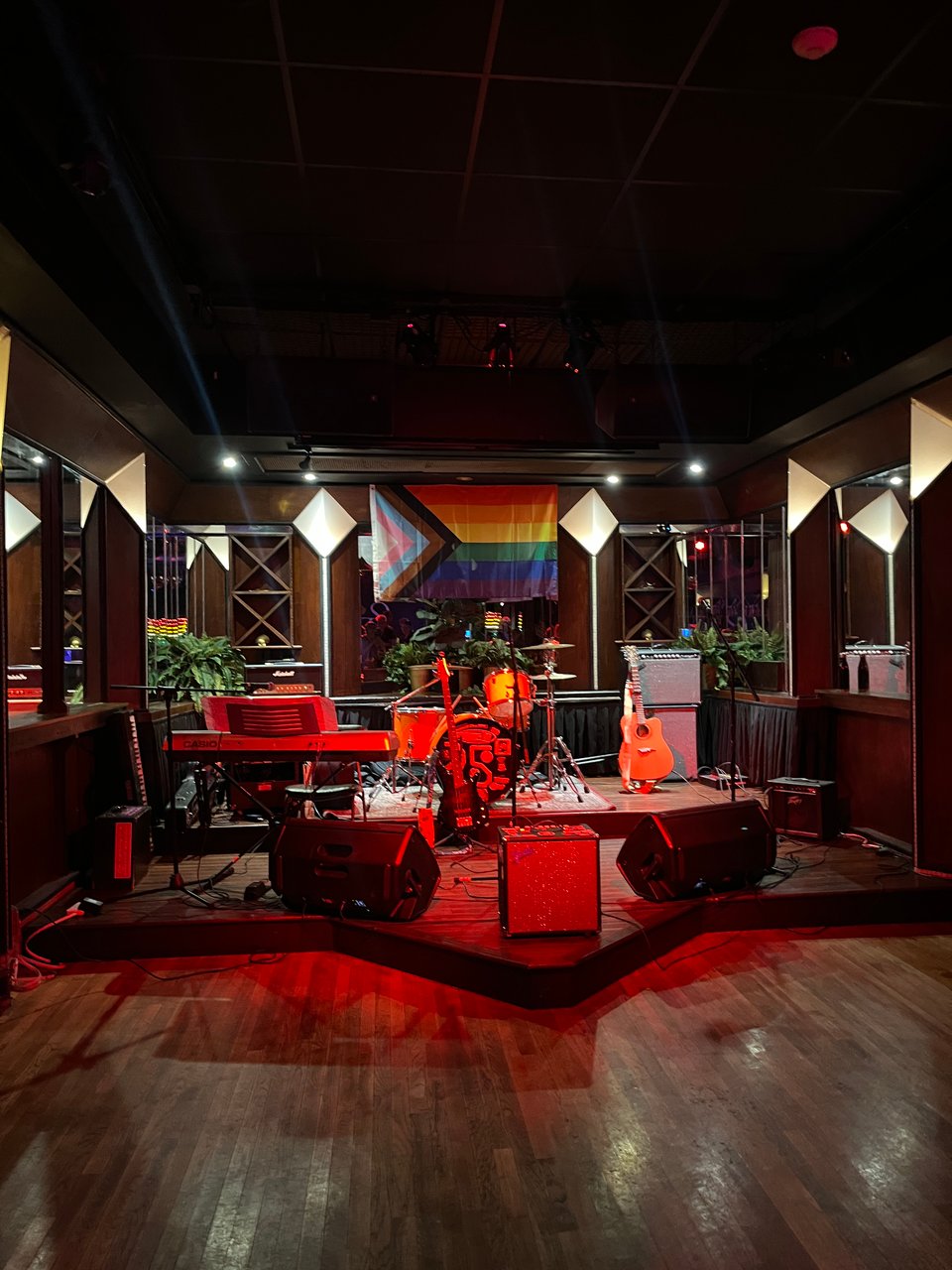 The Cardinal stage at night in a red-lit glow with instruments at the ready and a Pride flag in the background