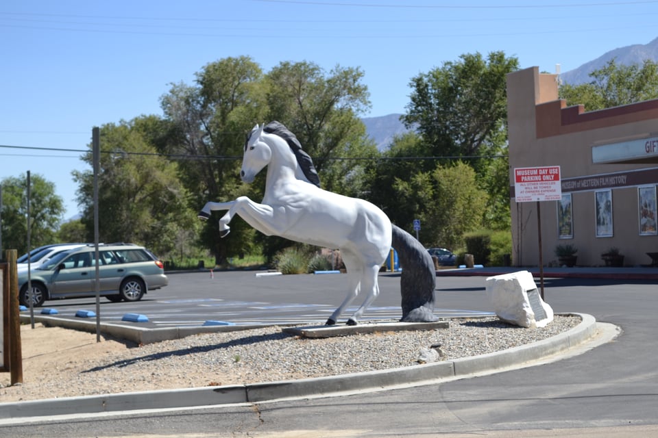 A statue of a (badly) rearing horse, clearly supported by its large tail. To the right is a small brick building and behind it is a parking lot.
