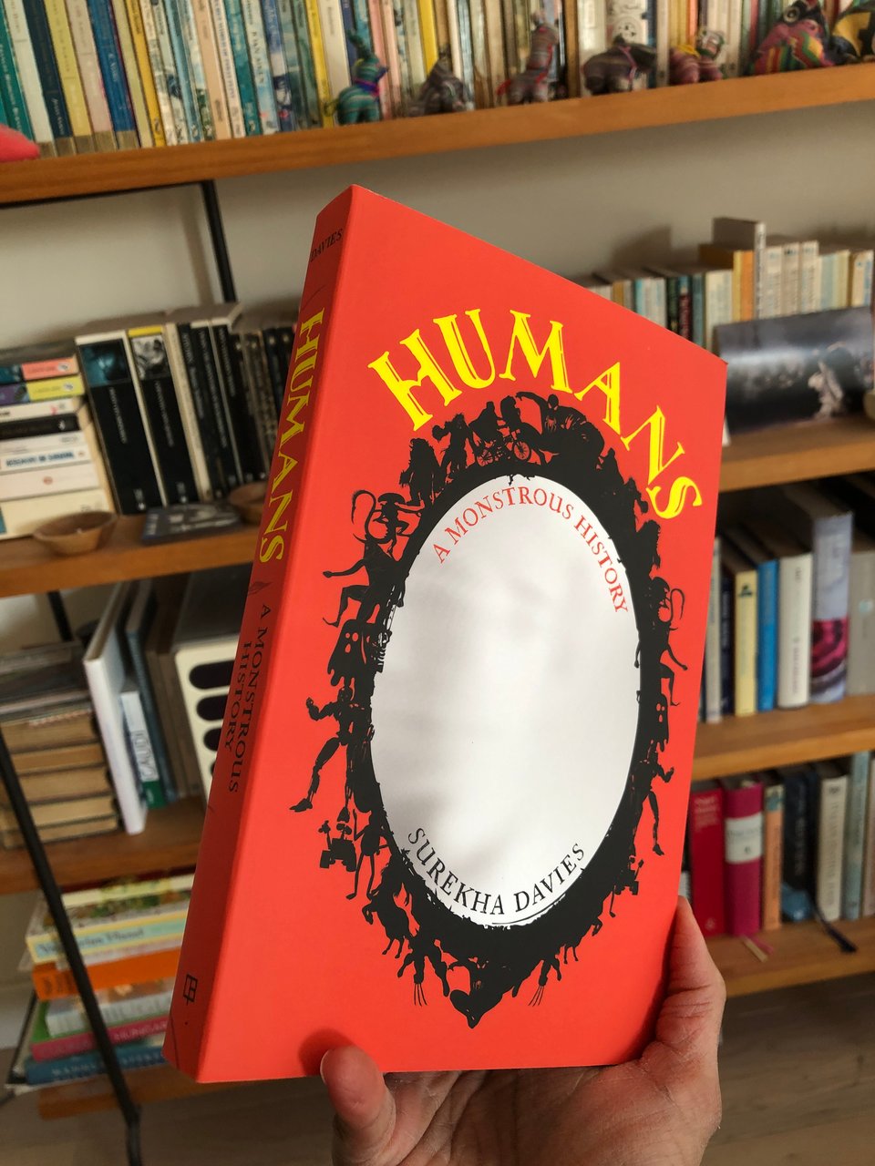 My right hand is holding up a paperback copy of HUMANS: A Monstrous History at an angle, in front of a bookcase brimming with brightly coloured books. The cover of HUMANS is mostly crimson, with bright yellow stylized lettering. A large, oval, whitish mirror with an elaborate black frame of SFF characters takes up the centre of the cover.