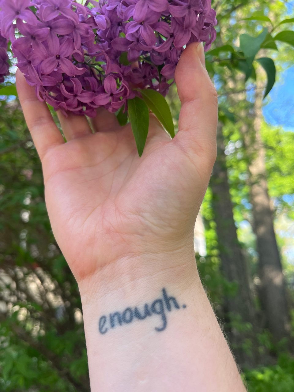 Nola's hand holding a dark purple lilac blossom, with her "enough" tattoo visible on her wrist